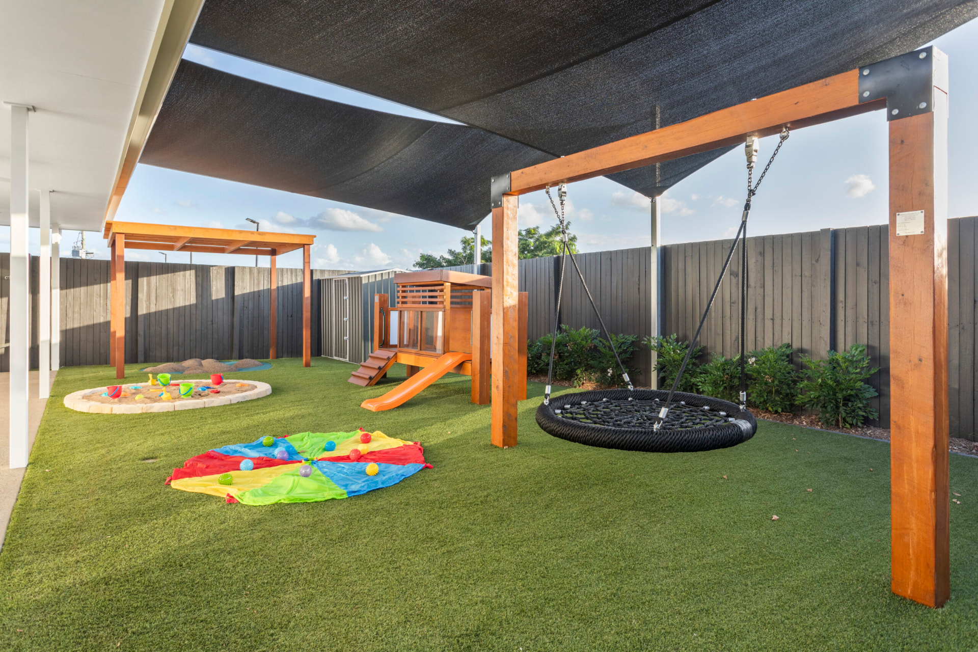 Shaded First5Years Calliope yard with a nest swing, sandpit, cubby and slide.