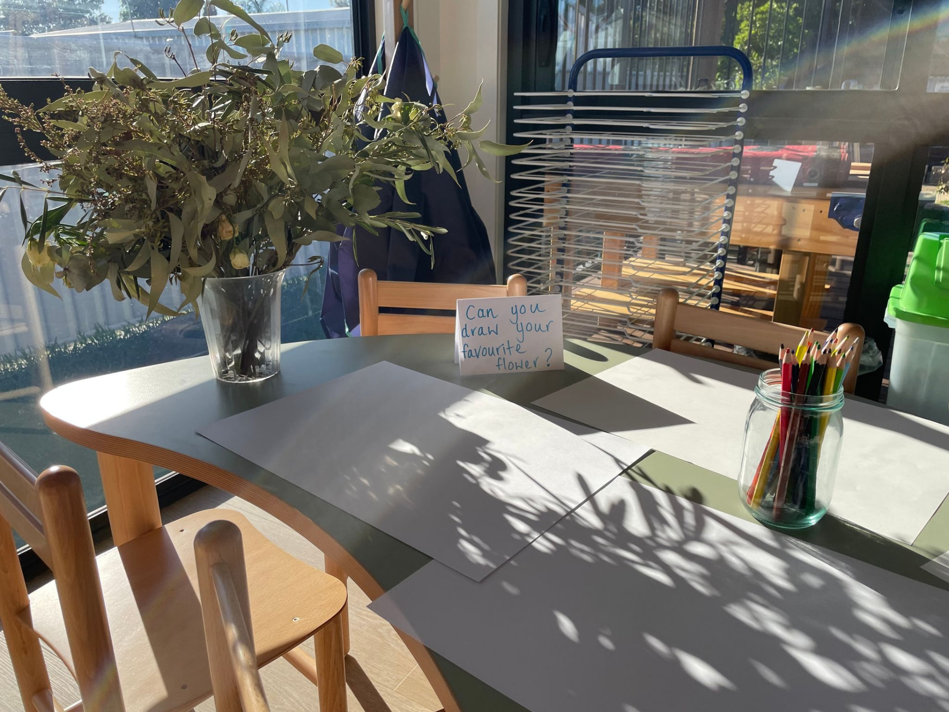 A table at First5Years Emerald set with paper, pencils and eucalyptus, with a handwritten card asking children to draw their favourite flower.
