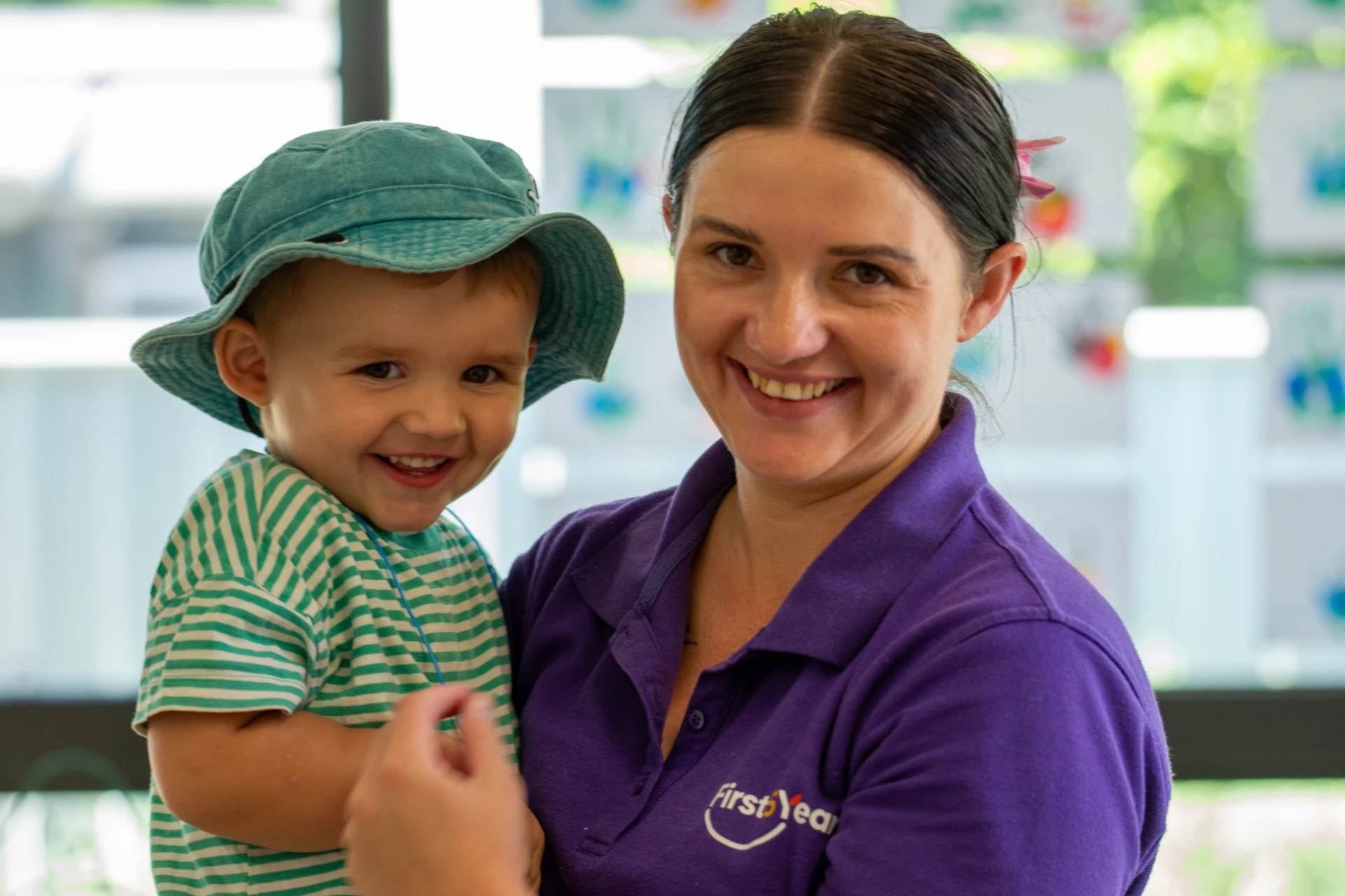 A First5Years educator in a purple uniform polo holding a smiling toddler in a sun hat.